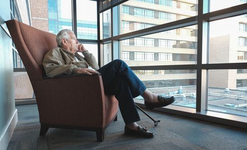 Side view of thoughtful man looking through window while sitting on armchair at home