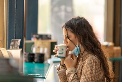 A beautiful teenage girl drinking coffee in bar wearing medical mask