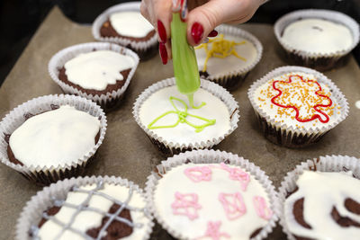 A woman squeezes colored frosting from a tube onto chocolate brown cupcakes covered white frosting.