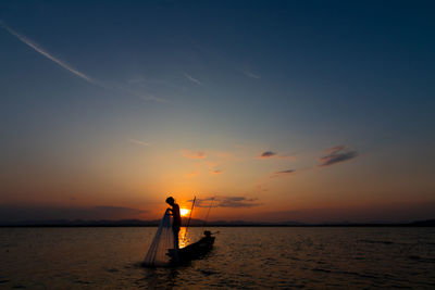 Silhouette man throwing fishing net in sea while standing in boat against sky during sunset