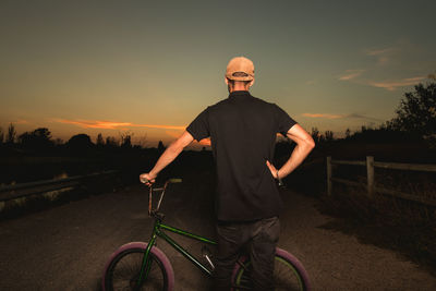 Rear view of young man with bicycle standing on road against sky during sunset