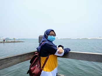 A girl wearing a blue hijab, mask and casual muslim t-shirt standing on the edge of the beach pier