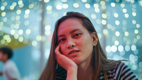 Close-up portrait of young woman against illuminated lights at night