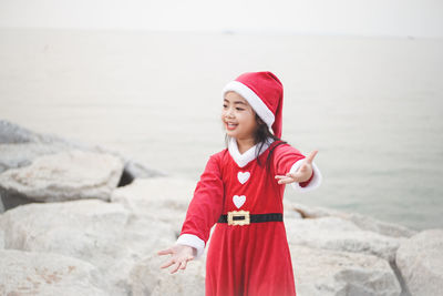 Smiling girl in santa claus costume standing on beach against sea and sky