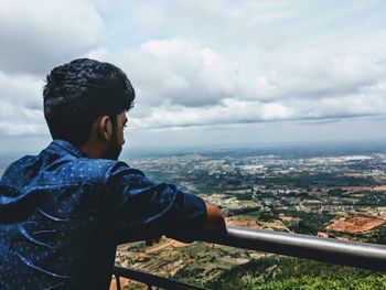 Rear view of man leaning on railing against cloudy sky