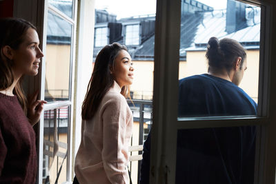 Smiling young friends looking through window while standing in balcony of rental apartment