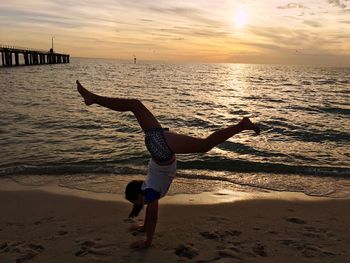 Full length of woman perching handstand at beach against sky during sunset