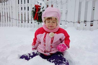 Girl in warm clothing sitting on snow covered field against fence