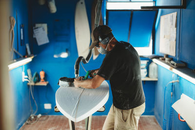 Worker in protective mask adjusting details surfboard in workshop