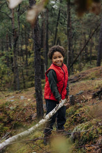 Smiling elementary boy picking up firewood while standing in forest