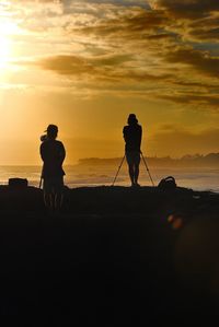 Rear view of people with tripod standing at sea shore during sunset