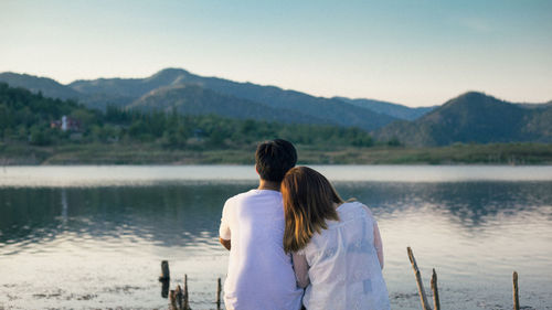 Rear view of couple sitting by lake against mountains