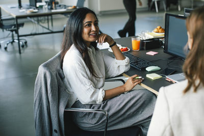 Side view of smiling businesswoman talking to colleague while sitting on chair in office