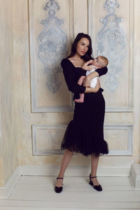 Mother in black dress holds a newborn boy in her arms at the window