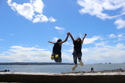 People holding hands while jumping at beach against sky