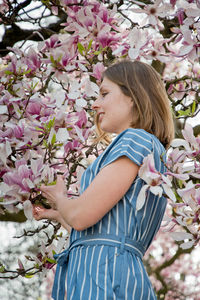 Blonde with pleasure inhales an aroman of magnolia flowers standing under a tree
