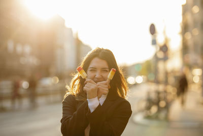 Portrait of young woman holding flavored ice while standing on road in city during sunset