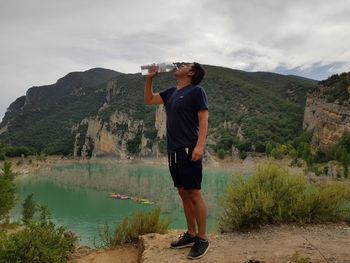 Man drinking water from bottle while standing on cliff by lake