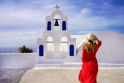 Woman with red dress and hat walking towards the bell of a traditional orthodox church in greece