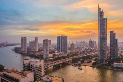 Modern buildings by river against sky during sunset