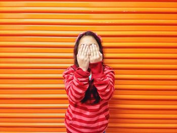 Front view of girl covering face with hands while standing against corrugated iron