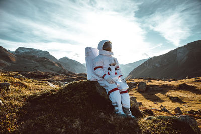 Astronaut sitting by rock on land against mountain