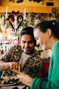 Smiling young man pointing at smart phone to woman sitting in restaurant during brunch party