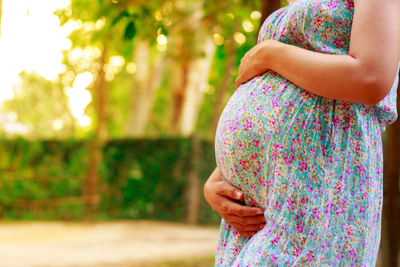 Midsection of pregnant woman touching abdomen while standing against trees