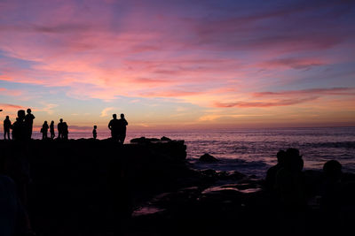 Silhouette people on rock by sea against sky during sunset