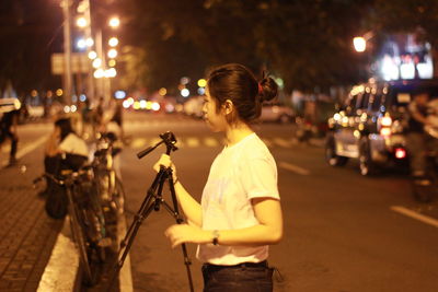 Young woman holding tripod on illuminated street at night