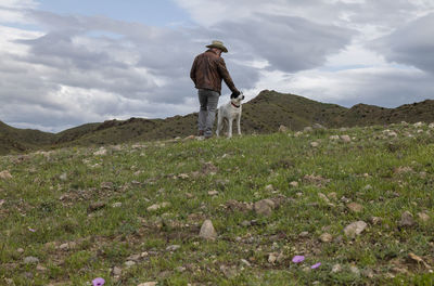 Adult man in cowboy hat and his dog standing on field against sky
