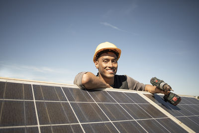 Portrait of smiling male engineer holding drill while leaning on solar panels at power station