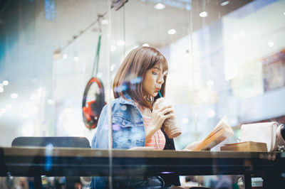 Young woman drinking cold coffee seen through window