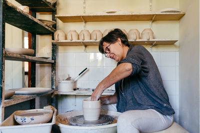 Middle aged craftswoman shaping ceramic vase on pottery wheel while working in craft studio