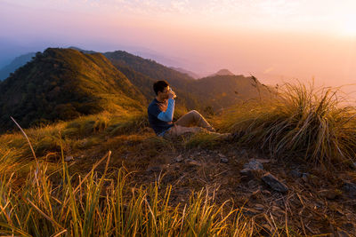 Side view of male hiker drinking while sitting on mountain against sky during sunset