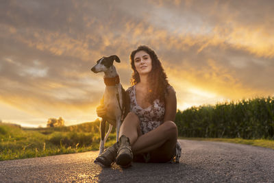 Portrait of woman with dog sitting on road against sky during sunset