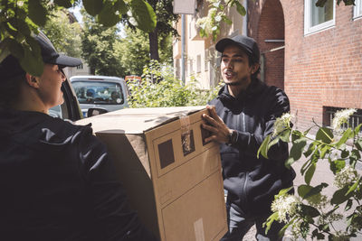 Smiling delivery colleagues talking and holding cardboard box near truck