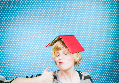 Close-up of woman with book on head gesturing thumbs up against wall