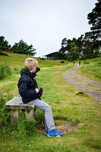 Boy holding binoculars while sitting on bench in grassy field at park