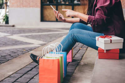 Low section of woman using mobile phone while sitting with shopping bags and gift boxes on footpath