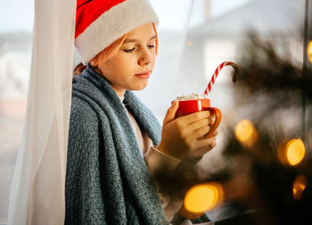 Young teen girl in santa hat having a cup of hot chocolate with marshmallow with christmas tree on