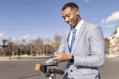 Smiling businessman unlocking electric push scooter through smart phone on sunny day