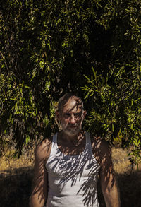 Adult man in white tank top and jeans standing with olive tree in summer