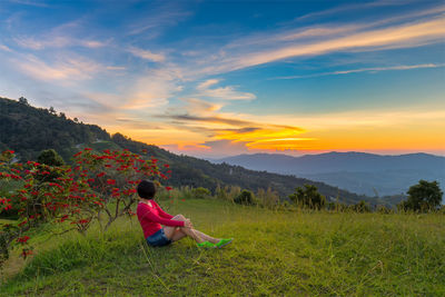 Woman sitting on grassy field against cloudy sky during sunset