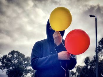 Front view of man holding red and yellow balloons against trees against sky.