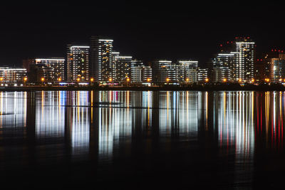 Illuminated buildings by river against sky at night