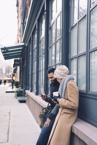 Couple using mobile phone while leaning by building in city