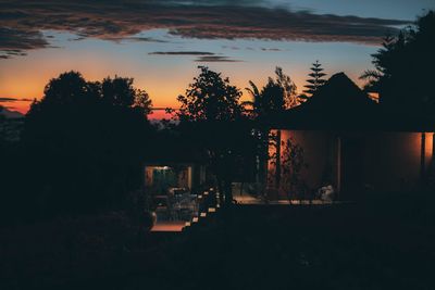 Silhouette trees by illuminated building against sky at sunset