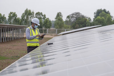 A young asian engineer wearing a helmet and white mask checks the cleanliness of solar panels.