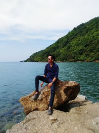 Full length portrait of young man sitting on rock by sea